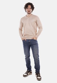 Maglione beige a scollo a V, in una texture lavorata a maglia, abbinato a jeans in denim blu. Sneakers casual marroni e bianche con un design testurizzato.