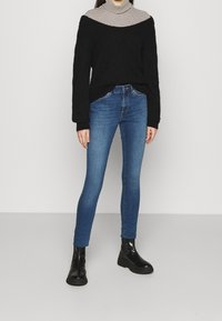 Suéter negro de gran tamaño con tejido texturizado, superpuesto a un cuello alto a rayas, combinado con jeans ajustados azules y botas negras de tobillo.