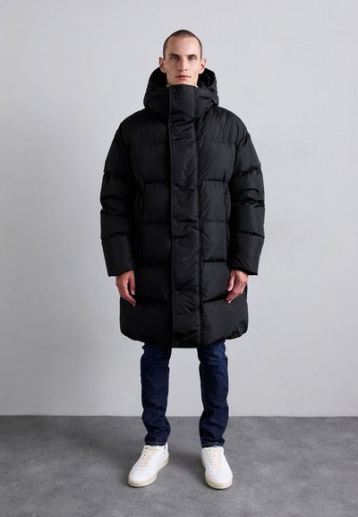 Homme debout portant un long manteau en duvet noir avec capuche, un jean bleu et des baskets blanches contre un mur gris clair uni.
