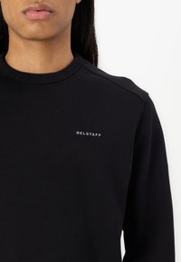 Black sweatshirt with a round neck, made of soft fabric. Features a small "BELSTAFF" logo in light gray on the chest. Smooth texture.