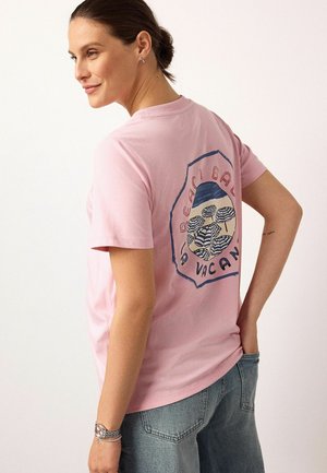 Woman wearing a pink T-shirt with a beach umbrella design and "Beach Ball Vacan" text on the back, paired with blue jeans.