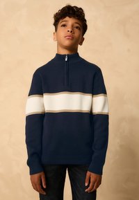 Laivastonsininen poolopusero puolivetoketjukauluksella, jossa valkoisia ja vaaleanruskeita vaakaraitoja. Karkea neulekangas ja pitkät hihat.