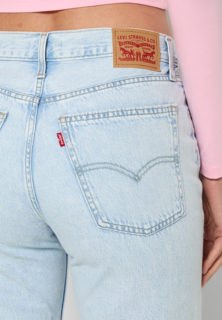 Jeans de mezclilla azul claro con dos bolsillos traseros, con un desvanecimiento sutil. Un parche de cuero marrón con el logo se encuentra en la cintura. Etiqueta roja en el bolsillo.
