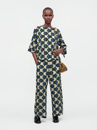 Femme portant un haut et un pantalon larges assortis à motif géométrique vert, bleu et beige, tenant un sac à main tressé marron avec une bandoulière chaîne.