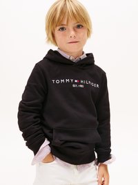 Melnā kapuce ar „TOMMY HILFIGER” logotipu un „EST. 1985.”. Uzlikta virs viegli svītrainas pogājamas krekla un baltām biksēm.