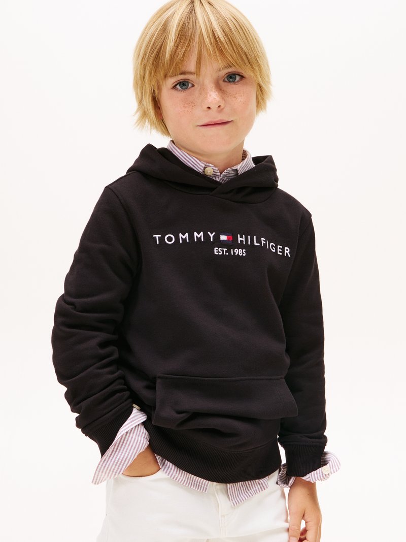 Melnā kapuce ar „TOMMY HILFIGER” logotipu un „EST. 1985.”. Uzlikta virs viegli svītrainas pogājamas krekla un baltām biksēm.