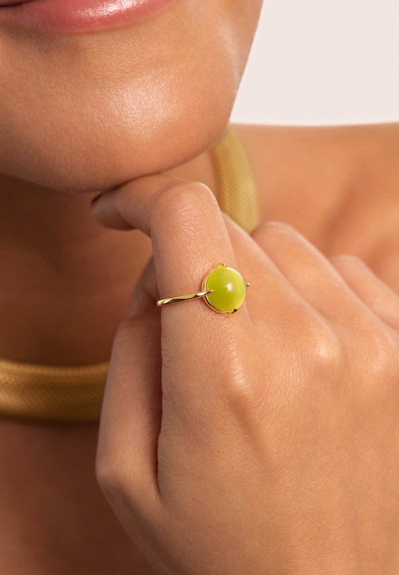 Goldring mit einem runden, durchsichtigen gelben Stein, der auf einem glatten Band sitzt, getragen an einer Hand in der Nähe des Kinns, mit einer goldfarbenen Netz-Halskette in der Nähe.