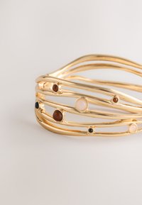 Gold-tone cuff bracelet with wavy metal strands and round gemstones in brown, black, and pale pink, featuring a textured finish.