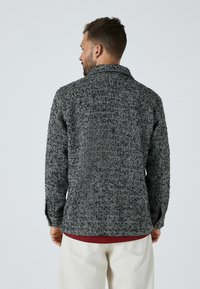 Kabát s herringbone vzorom v čierno-bielej kombinácii, vyrobený z texturovanej látky, s golierom a vo voľnom strihu, nosený cez vínovo červenú košeľu.