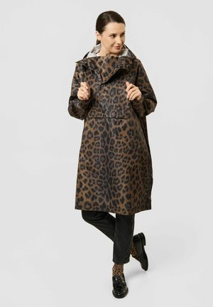 Femme portant un imperméable marron à motif léopard, chaussettes assorties, pantalon noir et chaussures noires, debout avec un pied croisé devant.