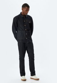 Chaqueta de mezclilla oscura con botones, bolsillos frontales con parche y pantalones de mezclilla oscura a juego. El modelo lleva zapatillas de color claro.