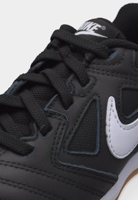 Μαύρο αθλητικό παπούτσι Nike με υφασμένες μαύρες κορδέλες, πλέγμα σε πάνελ, λευκό ραμμένο λογότυπο Nike swoosh και μαύρη γλώσσα με λευκό λογότυπο Nike.