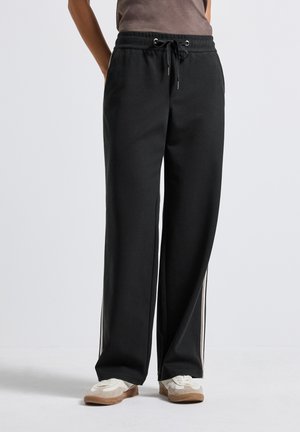 Personne portant un pantalon large noir avec des rayures latérales et des baskets blanches, se tenant devant un fond clair.