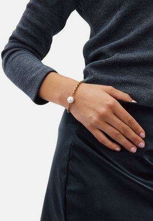 Bracciale a catena d'oro con un grande perno di perla come pezzo centrale, indossato su un polso con tonalità di pelle chiara, abbinato a una gonna nera e un maglione grigio.