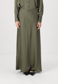 Jupe maxi en satin vert olive avec une texture lisse, dotée d'une fente sur le côté. Associée à des chaussures noires à bouts pointus.