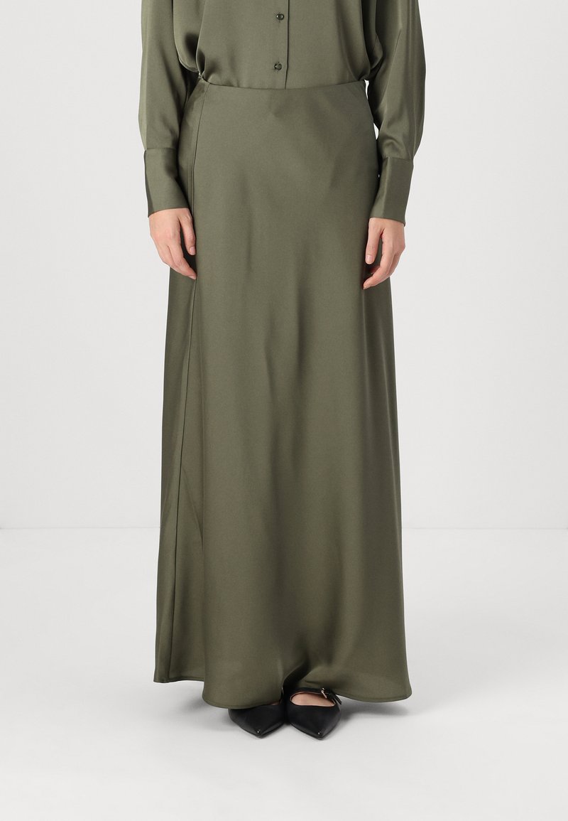 Jupe maxi en satin vert olive avec une texture lisse, dotée d'une fente sur le côté. Associée à des chaussures noires à bouts pointus.