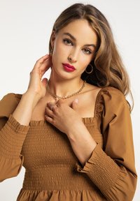 Hnědá smockovaná šaty s nafouknutými rukávy, s obdélníkovým výstřihem. Model má na sobě zlaté šperky a vlnité vlasy. Viditelná měkká textilie.