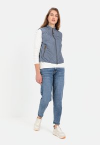 Grau gesteppte Weste mit hohem Kragen, Frontreißverschluss und zwei seitlichen Taschen. Tragen über einem weißen Langarmshirt, kombiniert mit blauen Jeans und Turnschuhen.