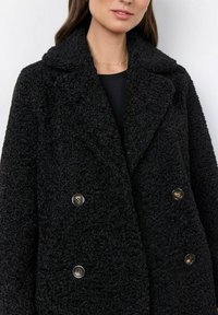 Abrigo negro de doble botonadura hecho de tela texturizada, con botones brillantes y un collar ancho, visible en un primer plano.