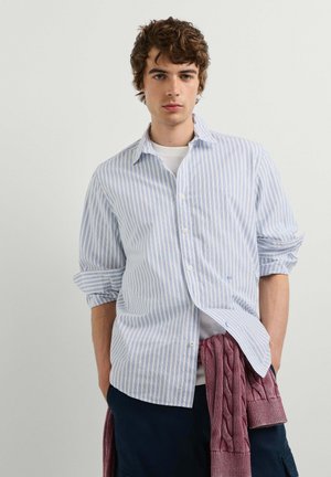 Jonge man met krullend haar, die een blauw-wit gestreept overhemd draagt over een wit T-shirt, met een bordeauxrode trui om zijn middel geknoopt.