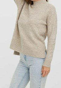 Maglione beige a maglia con colletto alto a coste, vestibilità ampia e tessuto testurizzato. Maniche lunghe e spacchi laterali all'orlo. Abbinato a jeans azzurri chiari.