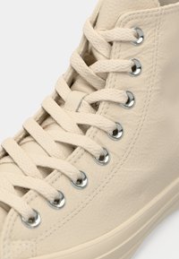 Baskets montantes beiges en tissu texturé, lacets beiges traversant des œillets métalliques argentés, avec des détails de couture visibles.