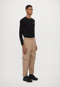Långärmad svart skjorta med beige logotyp; löst sittande beige cargobyxor med sidofickor; svarta sneakers.