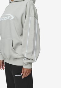 Sudadera con capucha gris claro con logo blanco, con bolsillo canguro, puños acanalados y franjas laterales blancas en contraste. Combinada con pantalones deportivos negros.