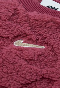 Bordový fleece sveter s textúrovaným materiálom, ktorý má zlatou vyšívané logo Nike a vínovo červený rebrovaný golier.