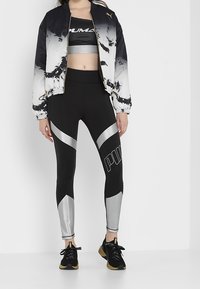 Krátká bombarová bunda PUMA v černé a bílé barvě s abstraktním designem, doplněná černými legínami s stříbrnými akcenty a logem, dokončená černými teniskami.