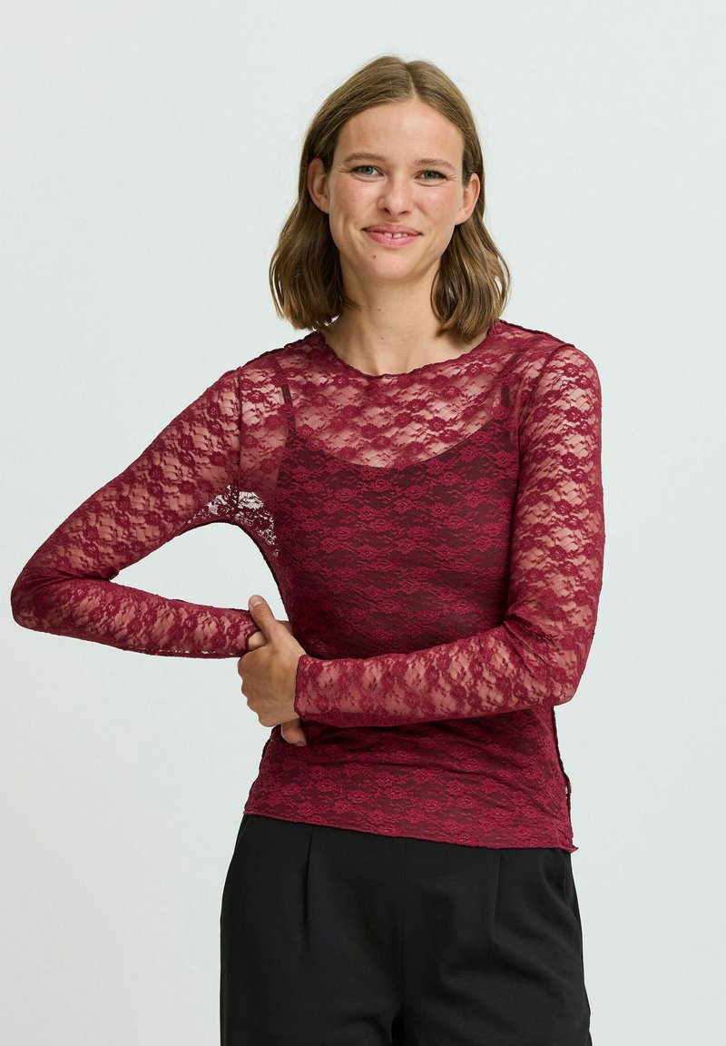 Top in pizzo bordeaux a maniche lunghe con motivo floreale, scollo a giro, e maniche trasparenti, abbinato a un fondo di colore uniforme.