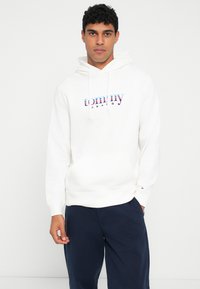 Λευκή φούτερ με μπροστινή τσέπη, κορδόνια και πολύχρωμο λογότυπο "tommy jeans". Ραμμένα μανίκια και κόψιμο. Φορεμένη πάνω από ναυτικά παντελόνια.