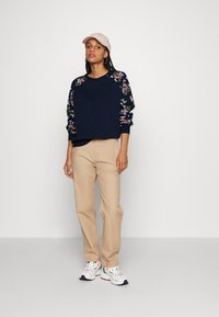 Navy sweatshirt with floral-embroidered sleeves, paired with beige pants and multicolored sneakers. Cap in pale pink completes the outfit.