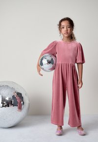 Combinaison rose côtelée avec manches courtes bouffantes, jambes larges et taille ajustée. L'enfant tient une boule disco réfléchissante et porte des chaussures roses scintillantes.