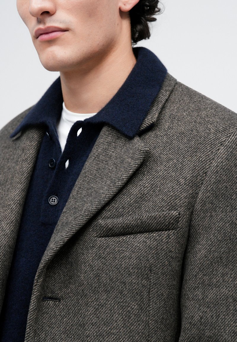 Homme portant un blazer en chevrons gris sur une chemise boutonnée bleu marine avec un sous-vêtement blanc, tourné légèrement à gauche sur un fond clair.