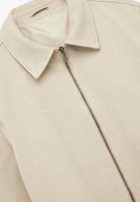 Beige zip-up jacket heeft een textuurstof, een spitse kraag en een ingewikkeld ritsdetail aan de voorkant. Gladde binnenvoering.