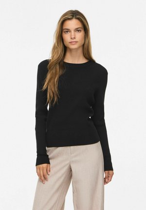 Ung kvinde med langt, lyst brunt hår iført en sort bluse med lange ærmer og beige bukser, stående foran en ensfarvet hvid baggrund.
