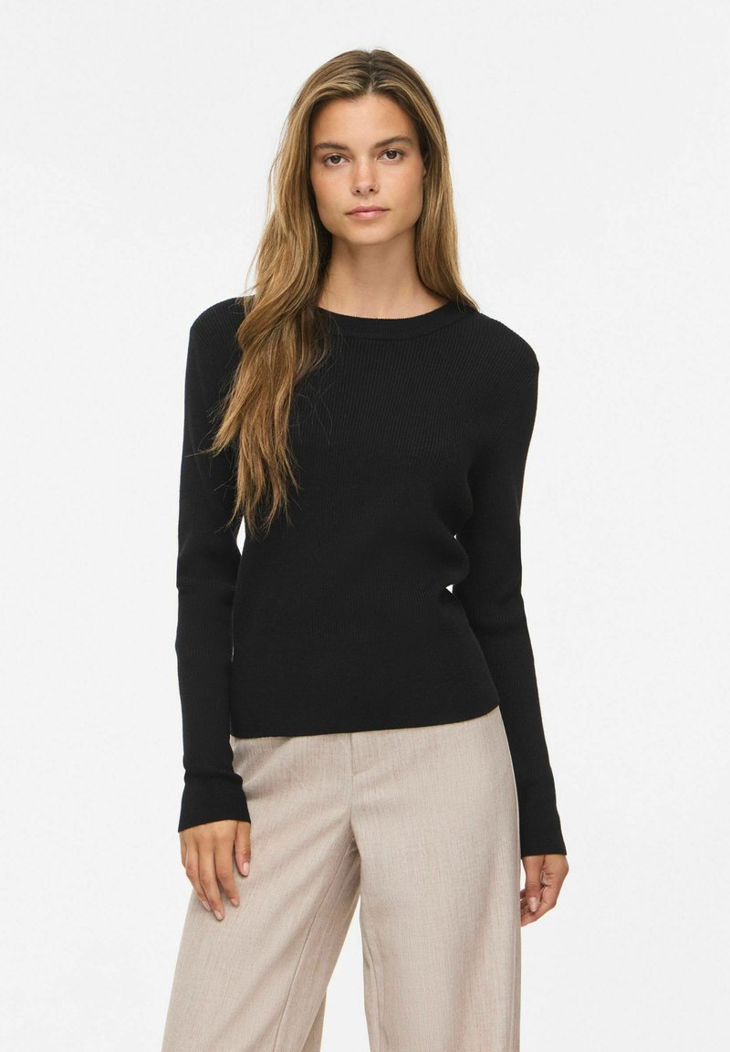 Jeune femme aux longs cheveux châtain clair portant un haut noir à manches longues et un pantalon beige, debout devant un fond blanc uni.