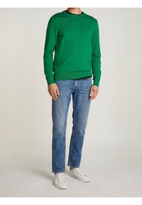 Suéter verde de punto con cuello redondo, combinado con vaqueros azules y zapatillas blancas. Tiene un pequeño logo en el pecho.