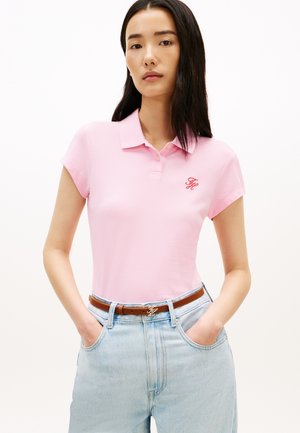 Žena s dugom tamnom kosom u ružičastoj polo majici kratkih rukava i svijetloplavim trapericama s smeđim remenom, rukama u džepovima, neutralna pozadina.