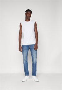 Λευκό αμάνικο μπλουζάκι, μπλε στενά τζιν και λευκά αθλητικά παπούτσια. Το μοντέλο έχει κοντά μαύρα μαλλιά και στέκεται μπροστά από ένα ουδέτερο φόντο.