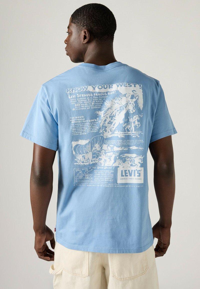 Mann trägt ein hellblaues Levi's T-Shirt mit weißen Western-Grafiken und Text auf dem Rücken, kombiniert mit hellen Shorts.