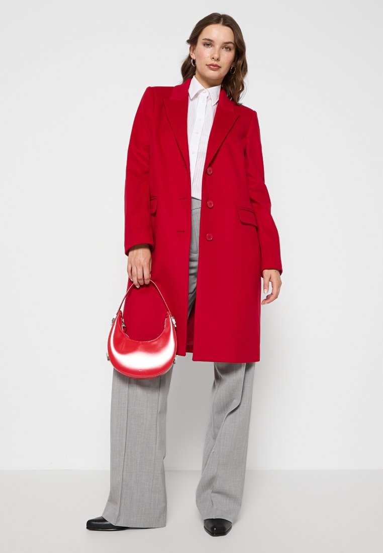 Cappotto rosso in misto lana con un design strutturato, chiusura a doppio bottone e tasche laterali; abbinato a pantaloni grigi a gamba larga e una borsa lucida rossa.