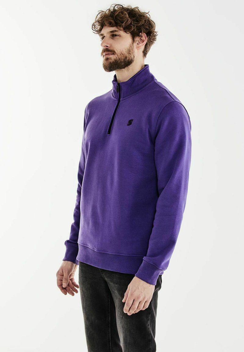 Purple pullover sweatshirt with a high collar, half-zip, and black logo on the chest; features ribbed cuffs and waistband, smooth texture.