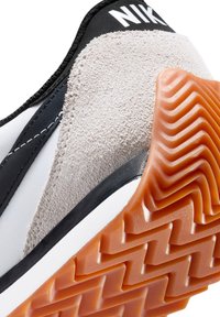Scarpa sportiva in bianco e nero con una tomaia in suede grigia testurizzata, suola in gomma con motivo a zigzag e dettaglio del logo nero sul tallone.