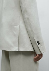 Helle beige Blazer mit Reverskragen, einer Schlitze hinten und einer Drei-Knopf-Manschette mit schwarzen Knöpfen. Glatte Textur.