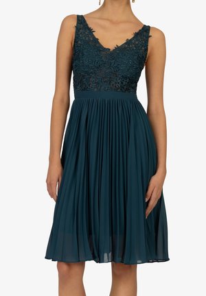 Robe sans manches vert sarcelle foncé avec corsage en dentelle florale et jupe plissée, portée par une personne avec des boucles d'oreilles dorées sur un fond uni.