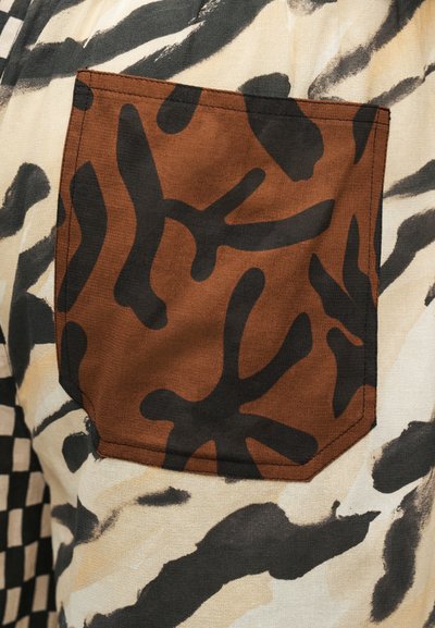 Poche en tissu marron avec des formes abstraites noires cousues sur un vêtement à imprimé animal crème et noir tacheté.