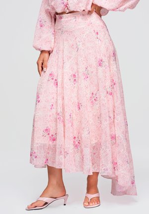 Jupe fluide rose à fleurs avec des détails en dentelle blanche associée à un haut assorti et des sandales roses à talons portées par une personne debout, main posée sur la hanche.