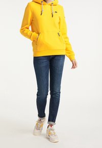 Sweat à capuche jaune avec poche avant, capuche à cordon et logo parapluie noir. Associé à un jean foncé et des baskets avec des accents jaunes.
